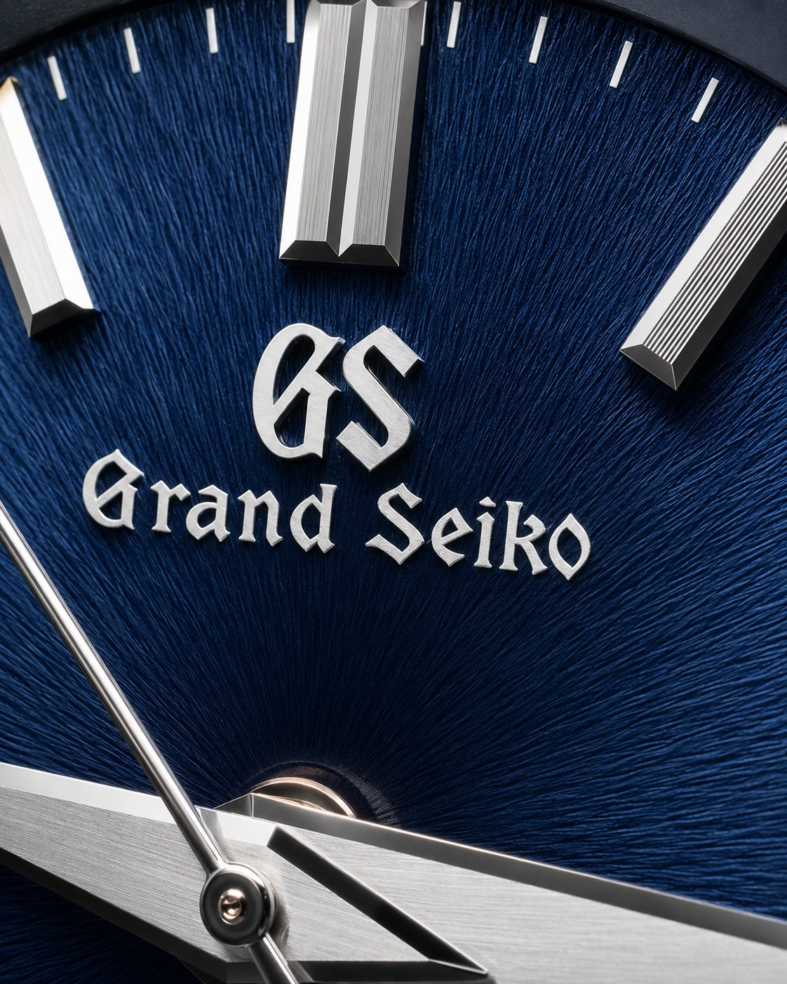 Close-up of a Grand Seiko watch dial with a deep blue textured background, silver baton indices, and the Grand Seiko logo and text in silver.
