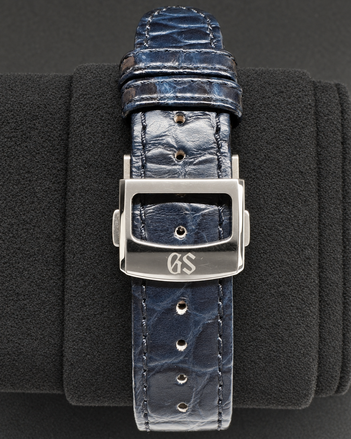 A close-up of a dark blue alligator leather watch strap with a polished silver-toned buckle engraved with the initials 'GS', displayed on a black textured cushion.