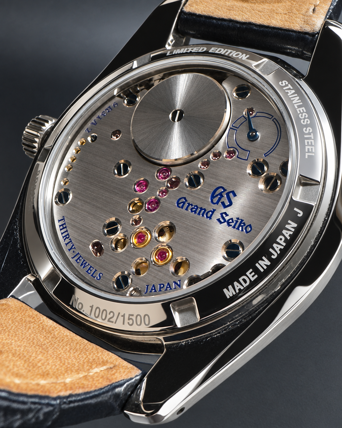 Close-up of the back of a Grand Seiko watch showing the stainless steel case, exposed mechanical movement with jewel bearings, and a dark leather strap on a dark studio background.