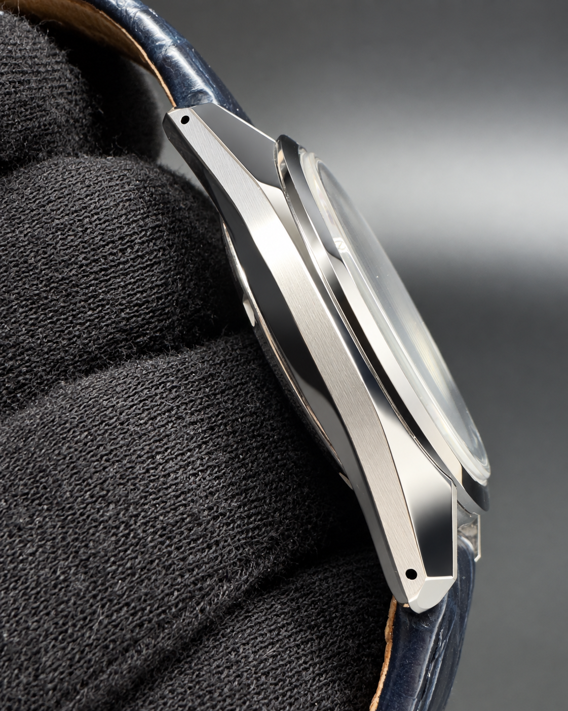 Close-up side profile of a polished silver-toned metal watch case with a dark blue leather strap, resting on a textured black fabric surface against a dark gray background.
