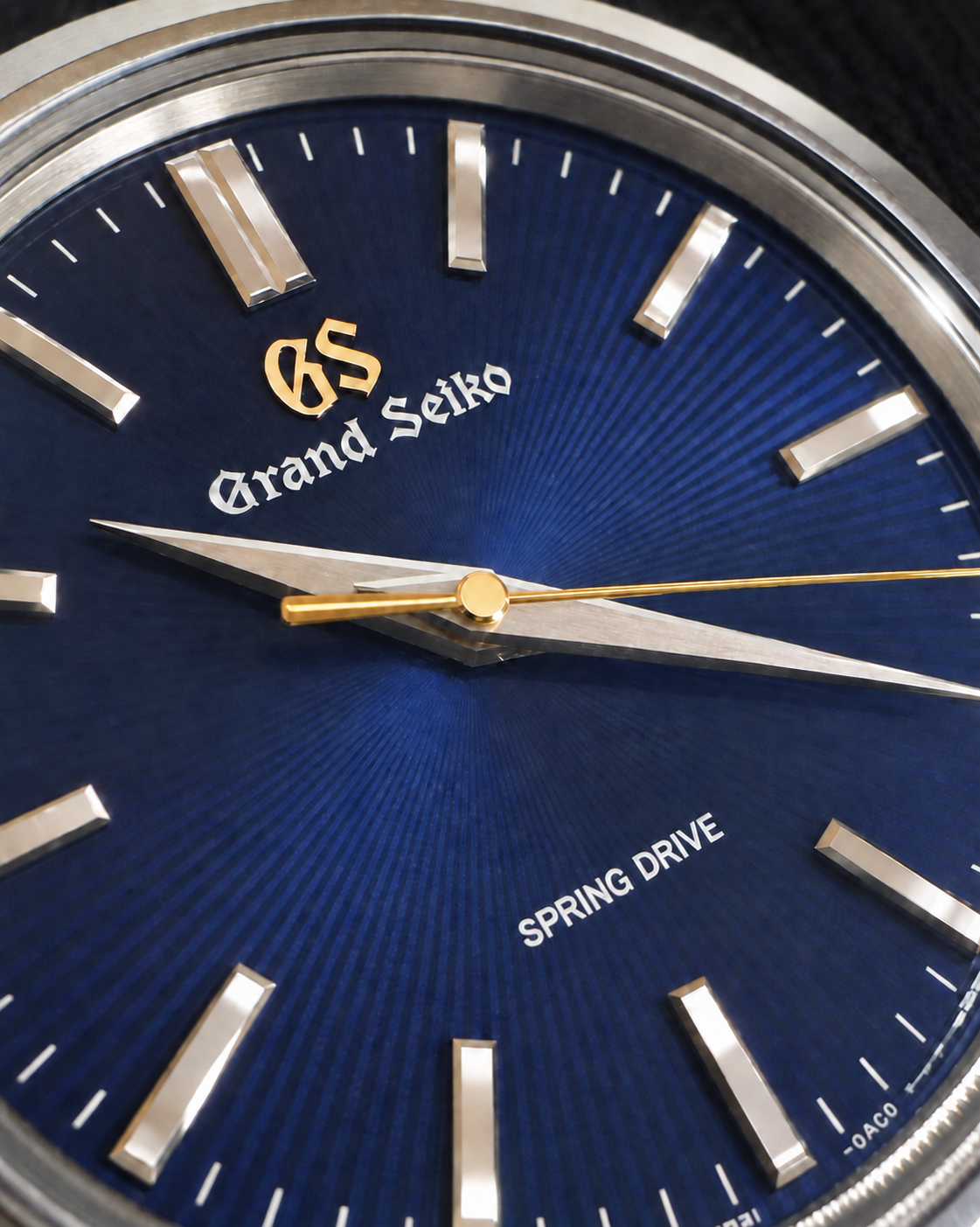 Close-up of a Grand Seiko watch dial with a deep blue sunburst face, silver baton hour markers, silver hands, and a thin gold seconds hand.