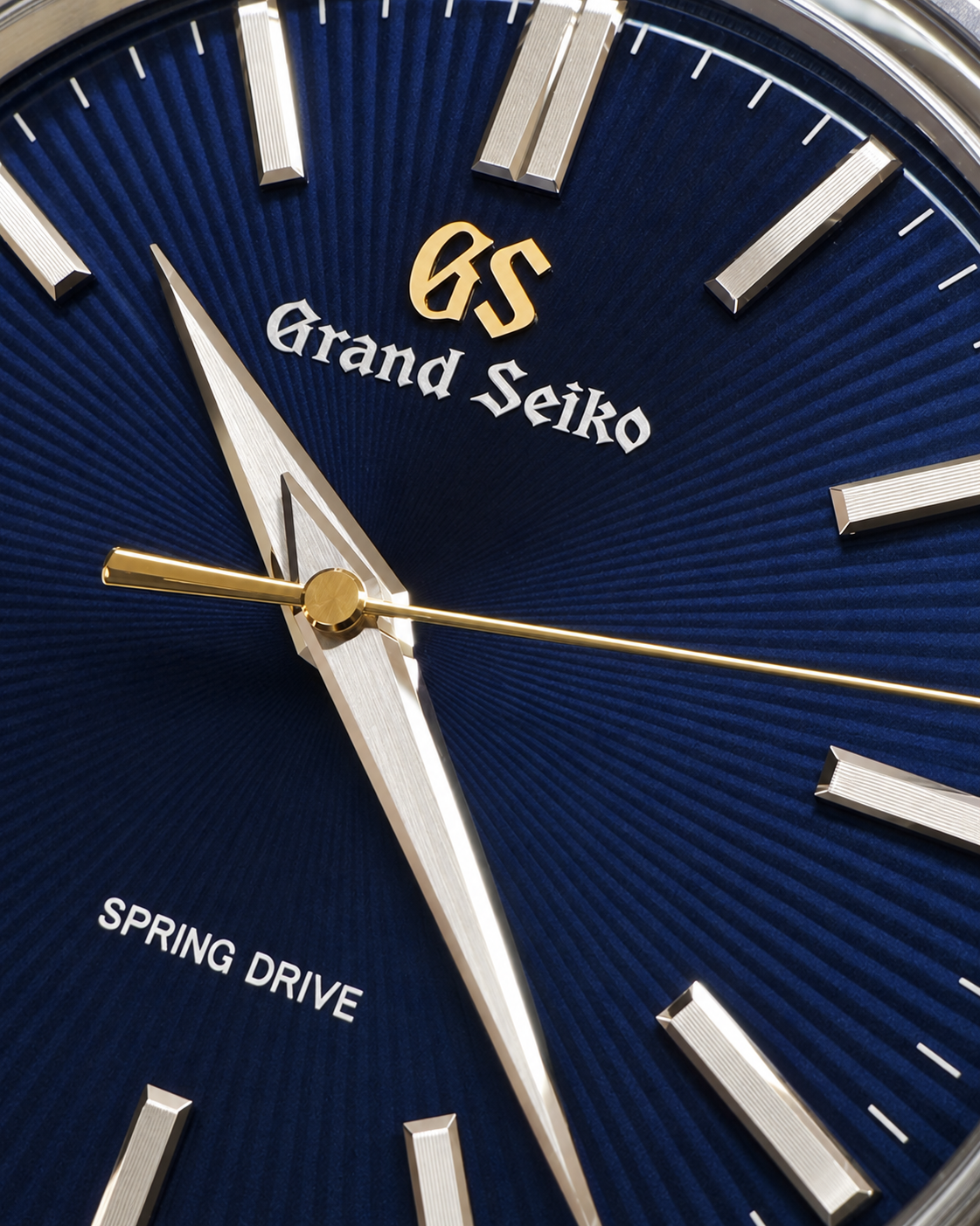 Close-up of a Grand Seiko watch dial with a deep blue sunburst background, silver stick indices, gold hands and center marker, and white brand text.