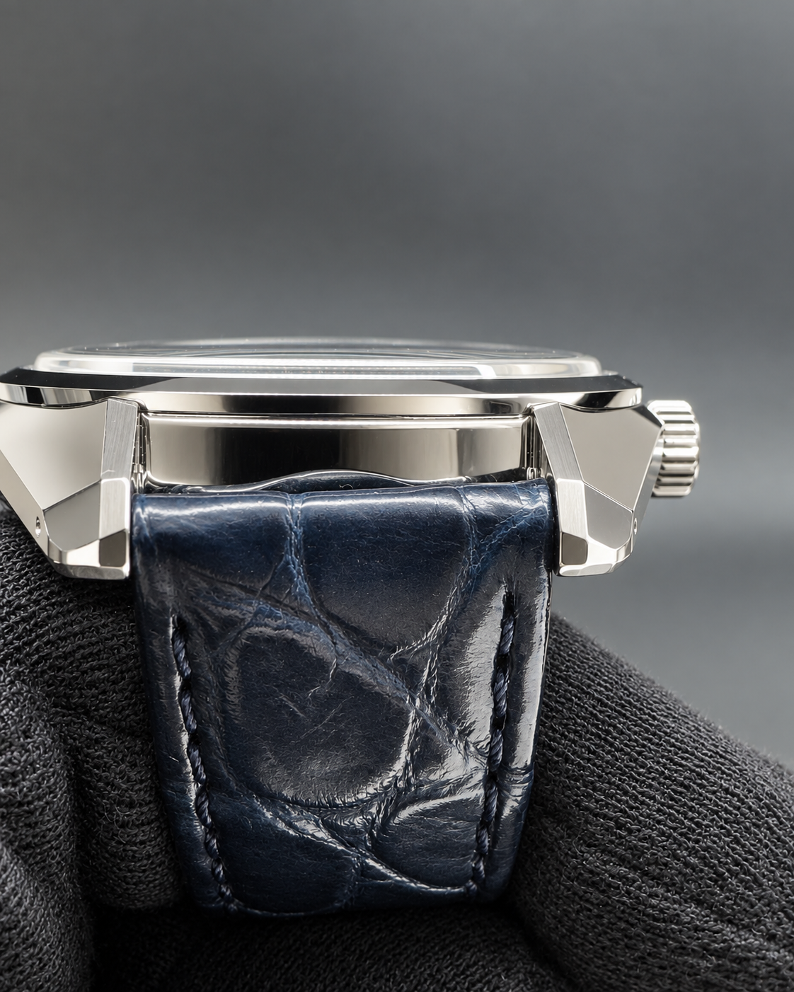 Close-up of a wristwatch with a silver-toned metal case and a dark blue alligator leather strap, resting on a dark textured surface against a gray background.