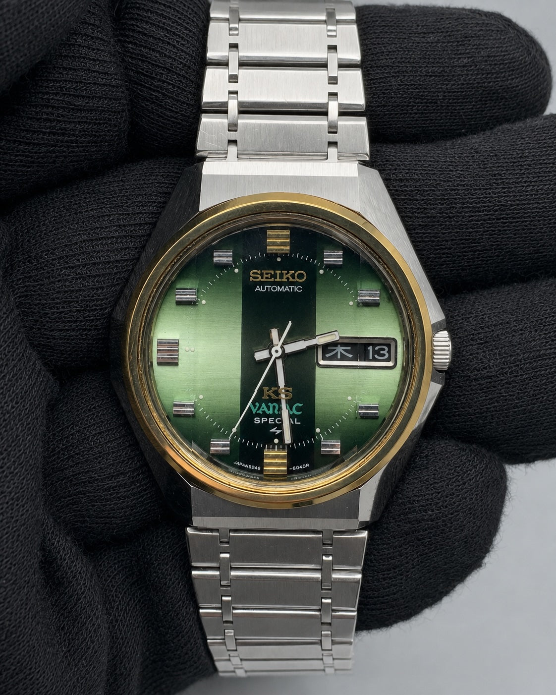 A Seiko KSC 8ATM Automatic watch with a green dial and a gold-tone bezel, held by a black-gloved hand against a neutral gray background.