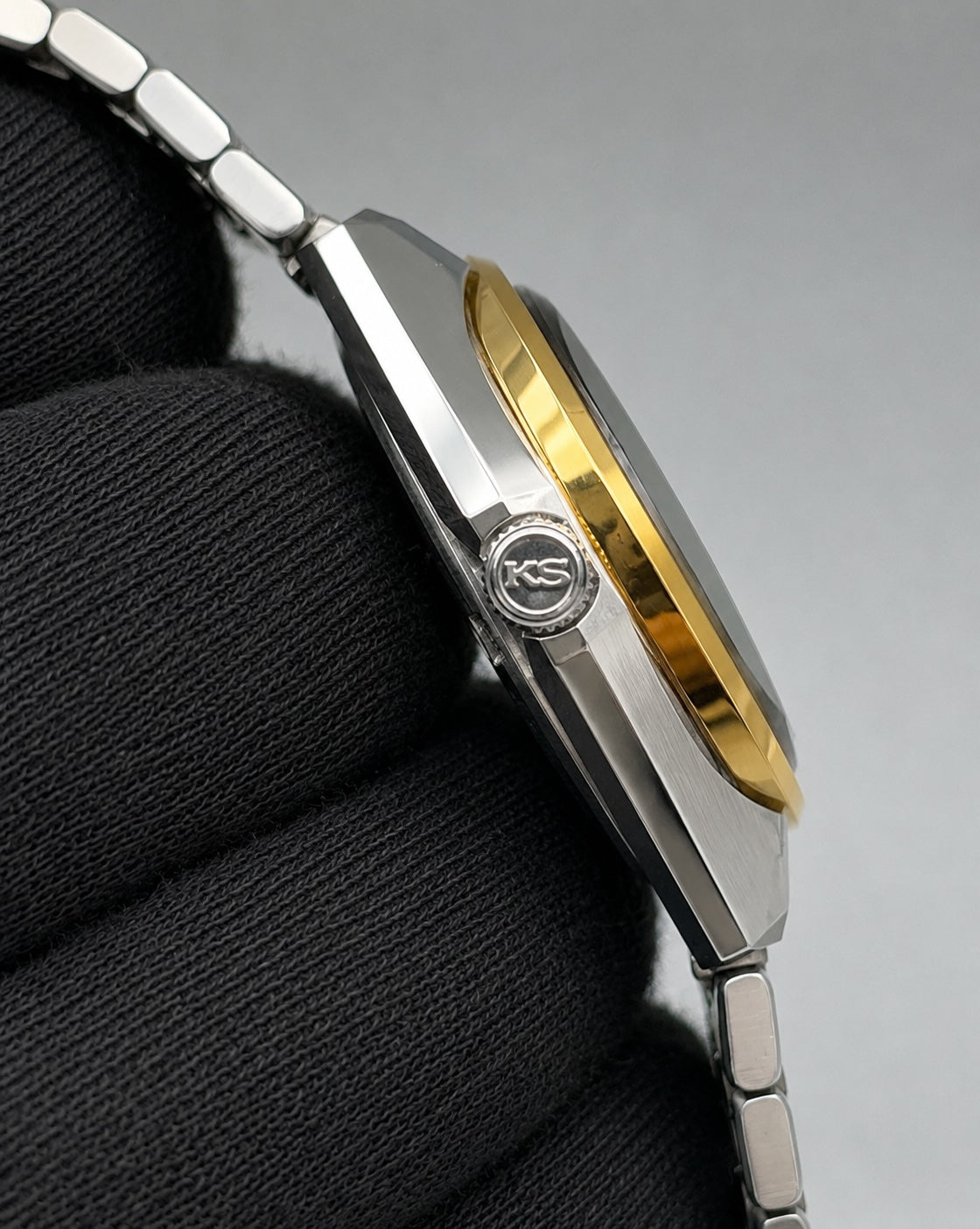 Close-up side view of a two-tone stainless steel and gold watch with a silver link bracelet, resting on a black textured surface against a gray background.