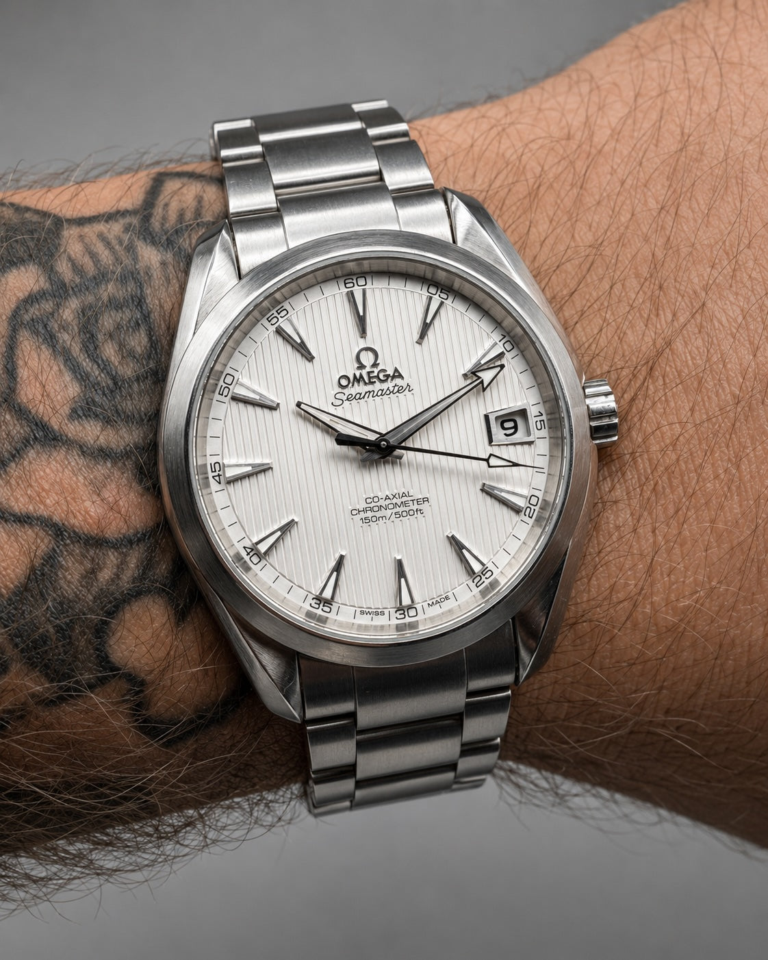 A close-up of an Omega x Cosmopolitan wristwatch with a silver stainless steel Oyster bracelet and a white dial, worn on a person's tattooed wrist.