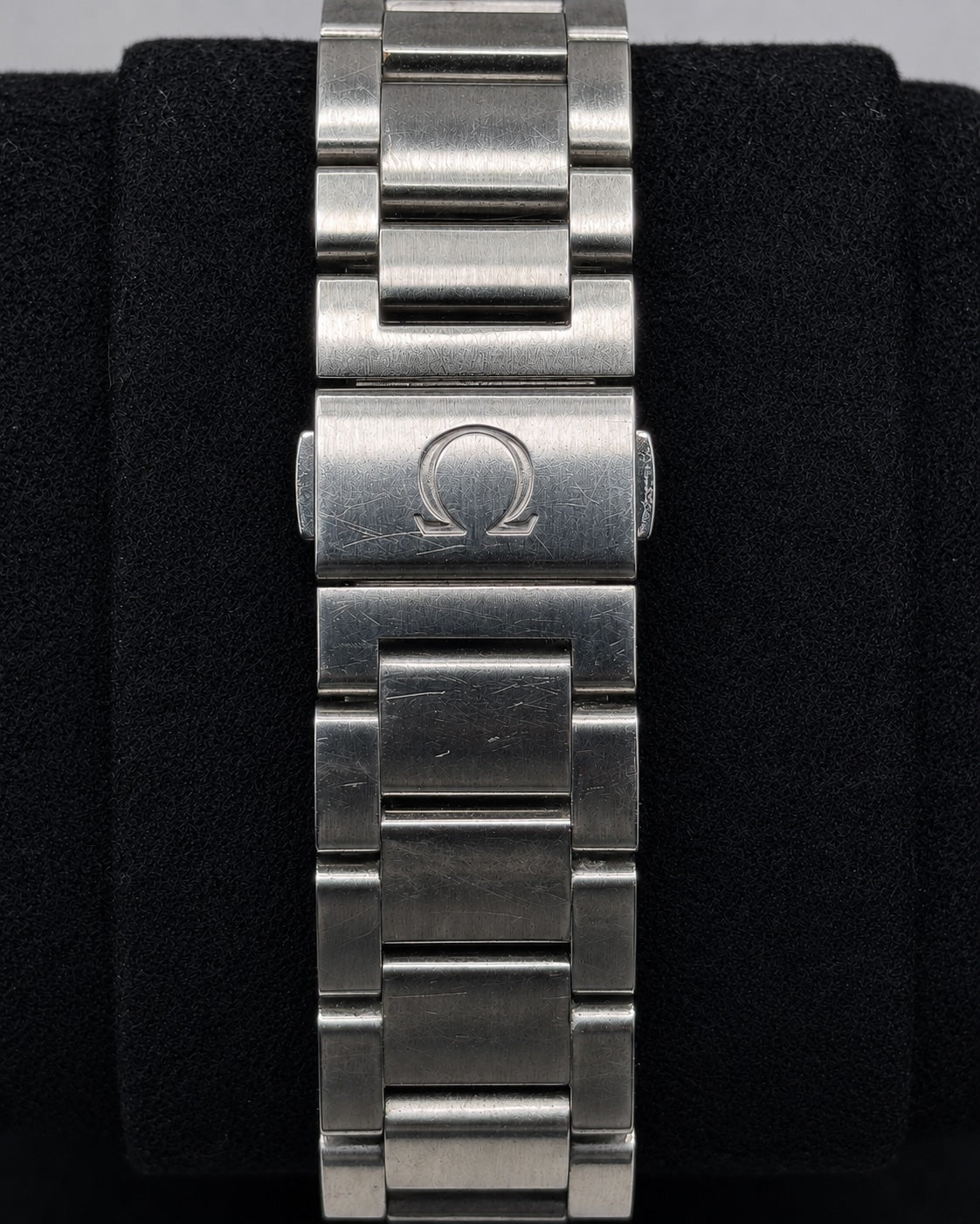A close-up of a stainless steel watch band clasp featuring the engraved Omega logo, displayed on a black velvet stand.
